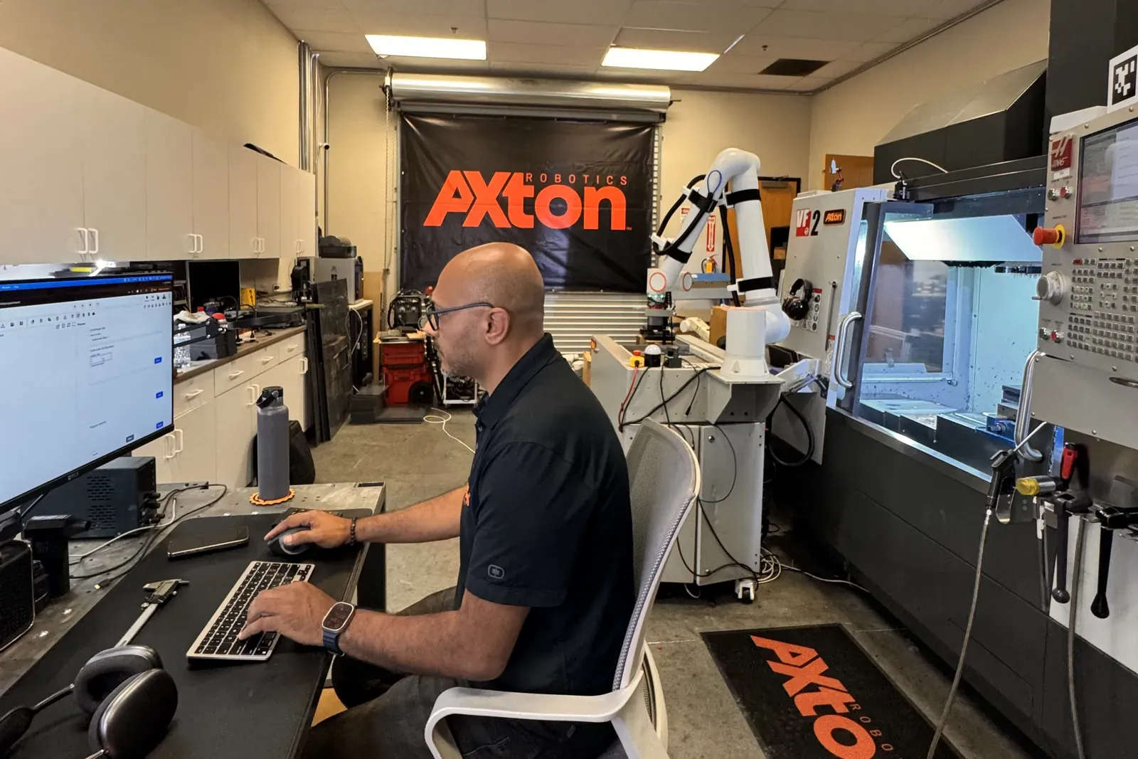 Michael Nessim programming at his desk, with the cobot cell and a Haas CNC mill beside him in the Axton Robotics lab